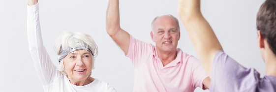 eine ältere Dame und ein älterer Herr sitzen mit ihrem Fitness-Trainer gegenüber, sie heben ihren Arm weit nach oben